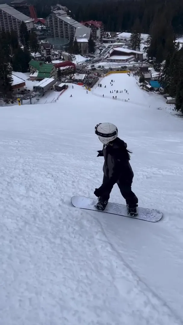 Child on a snowboard riding down a wide piste above the hotels and lift base in Borovets