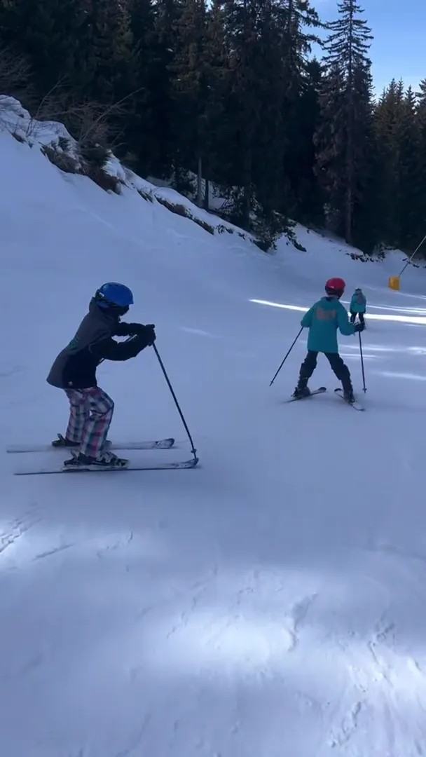 Two young skiers practicing turns on a shaded slope beside a pine forest