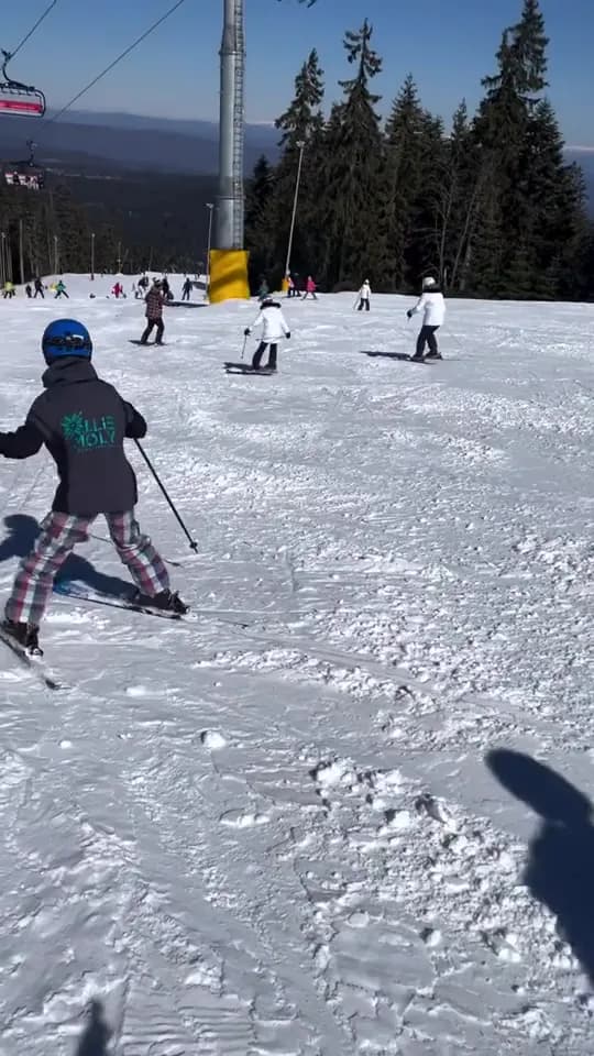 Child in a blue helmet skiing toward other young skiers under a chairlift