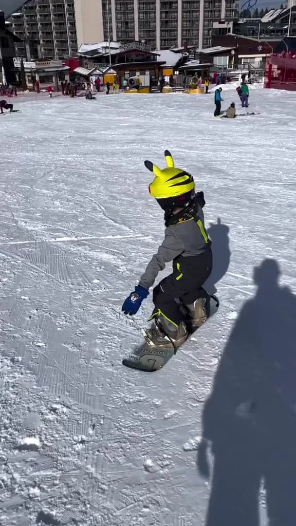Young snowboarder in a bright yellow Pikachu helmet riding across the Borovets base area