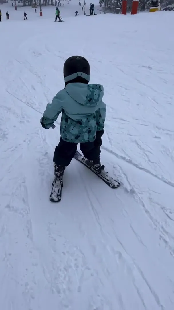 Small child in a mint-green jacket holding a snowplough position on a gentle beginner slope Rotata