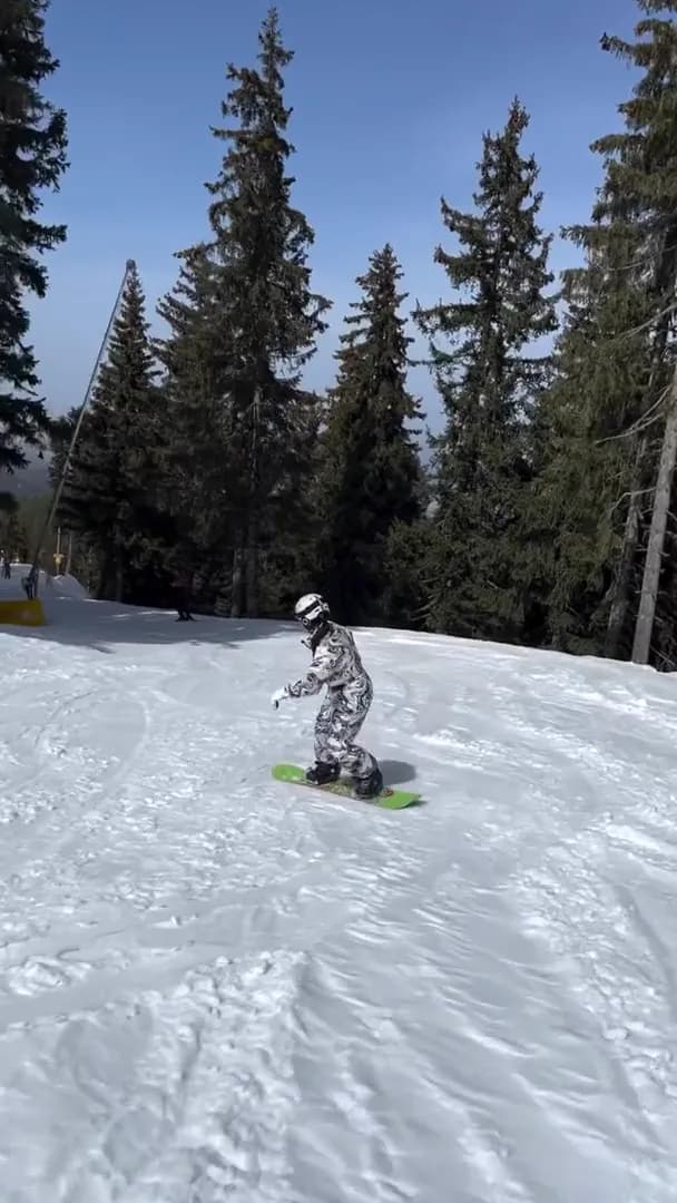 Snowboarder in white camouflage gear riding a groomed run between tall spruce trees